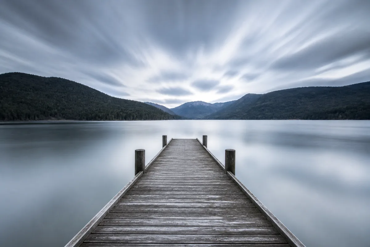 Jetée de bois s&rsquo;avançant sur un lac lissé, nuages filés convergeant au-dessus de l&rsquo;horizon