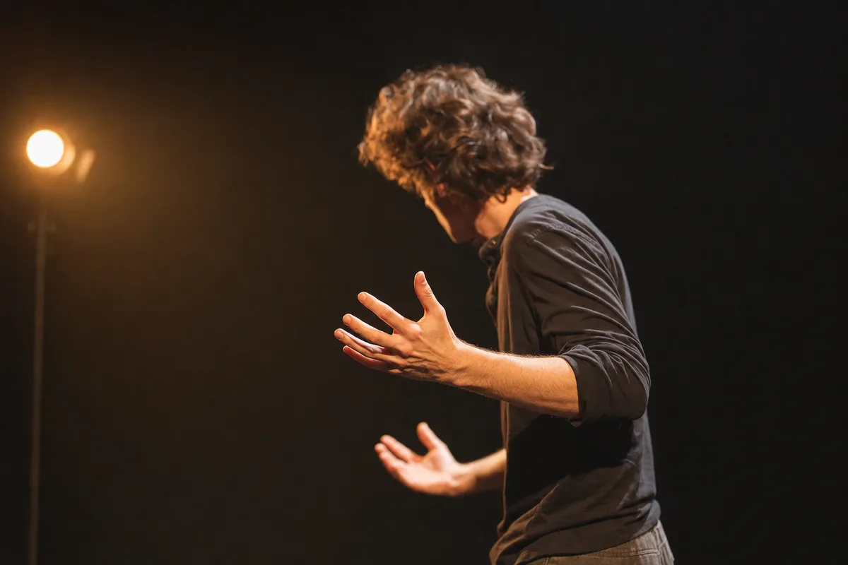Comédien sur une scène de théâtre sombre, geste expressif sous un projecteur latéral chaud